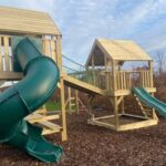 Wooden playground structure with a green spiral slide and bridge, set on mulch under a clear sky.