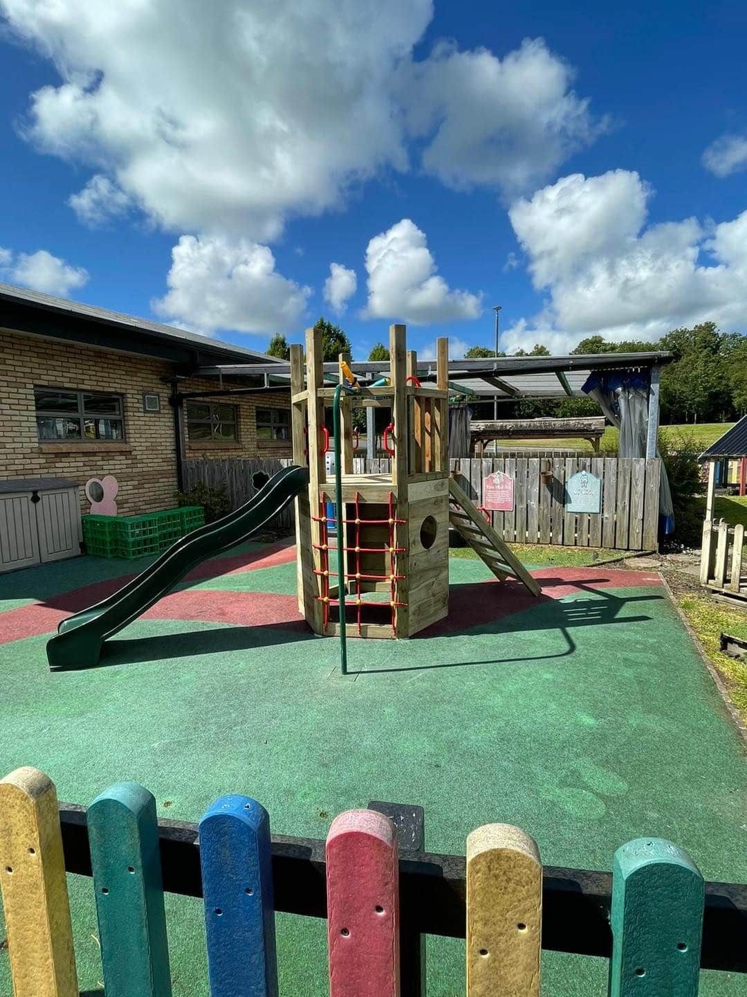 Playground with a wooden climbing structure, slide, and colorful padded surface under a blue sky with clouds.