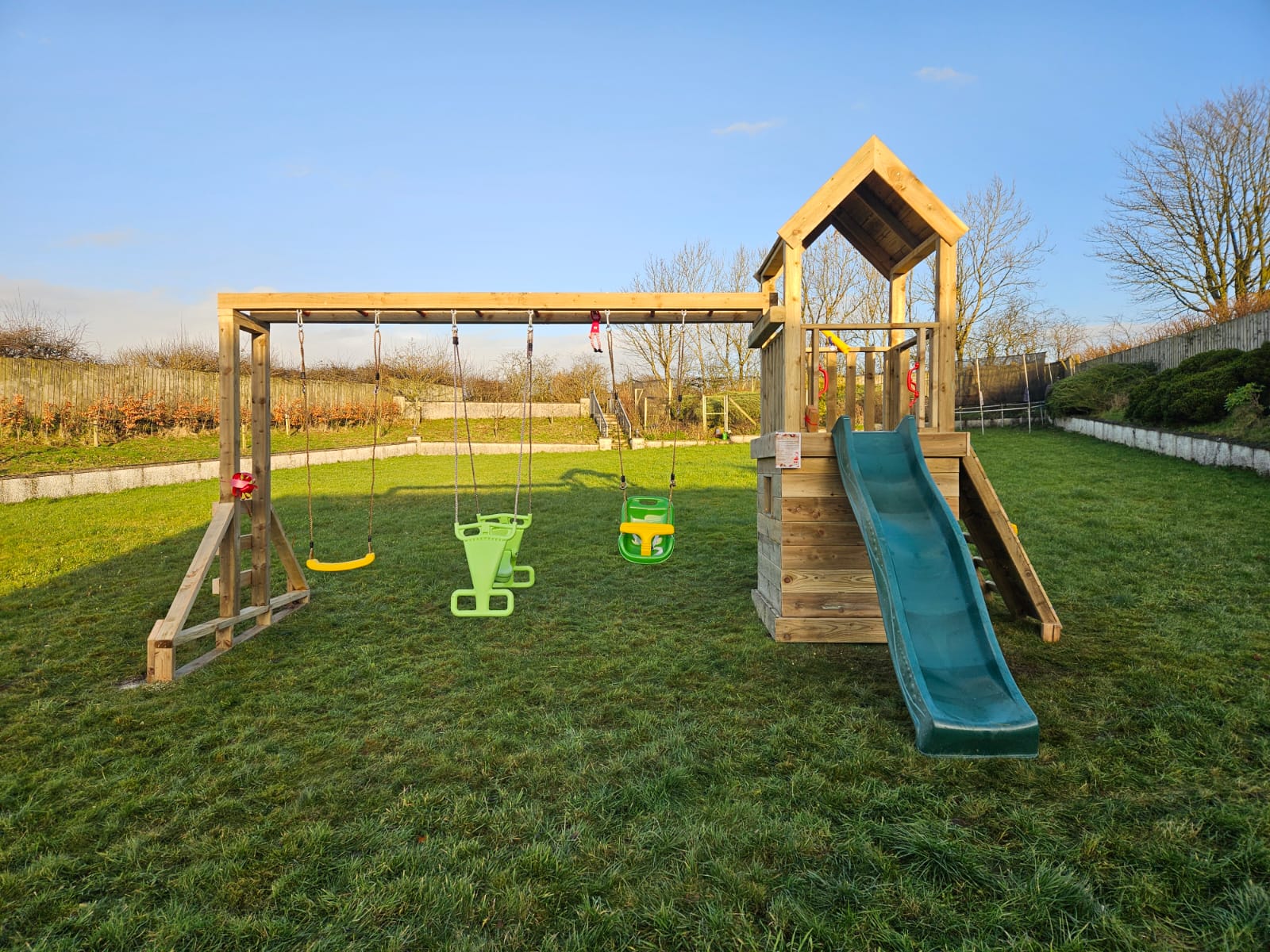 Wooden playground set with a slide and swings in a grassy backyard under a clear blue sky.