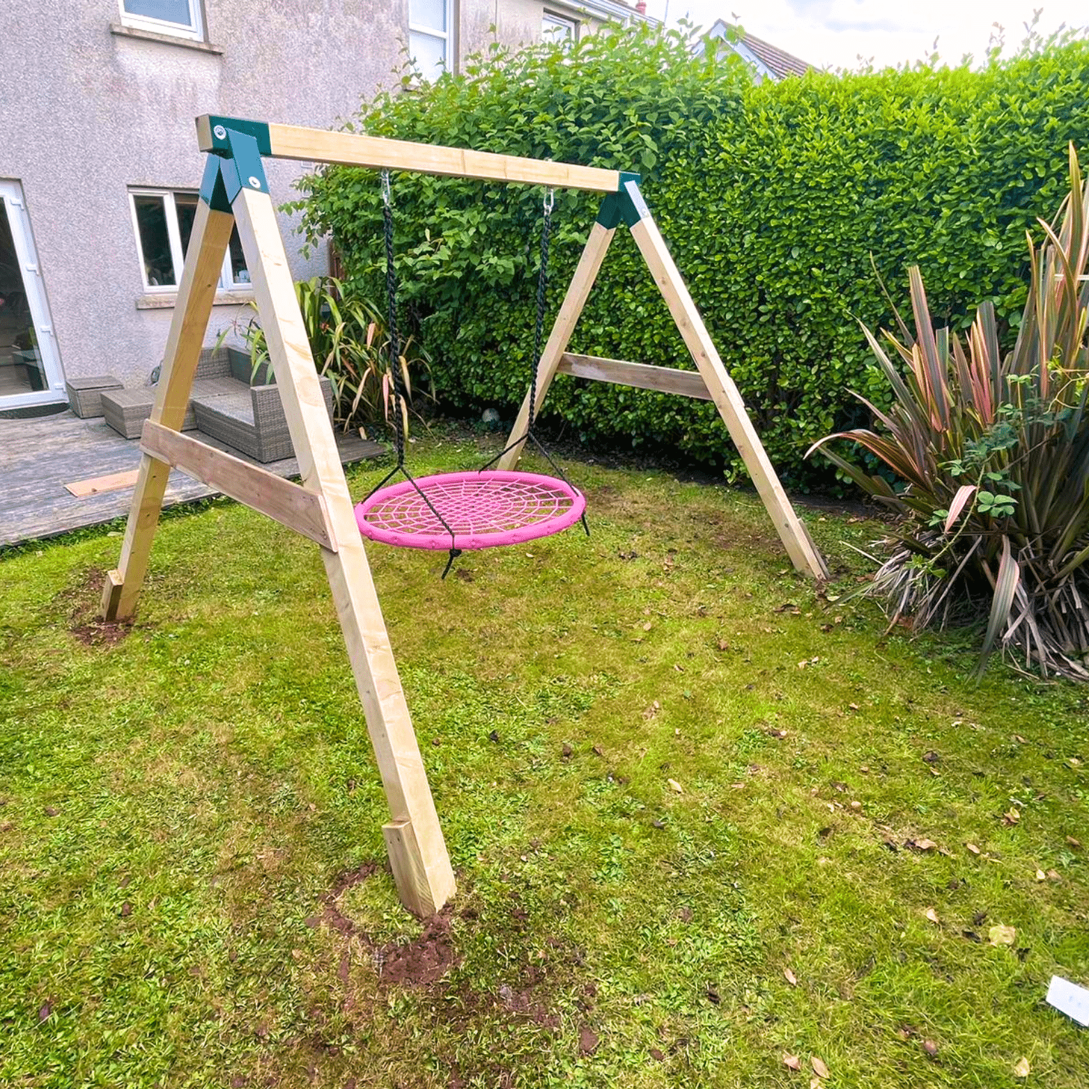 Pink circular swing on a wooden frame in a backyard with grass and bushes.