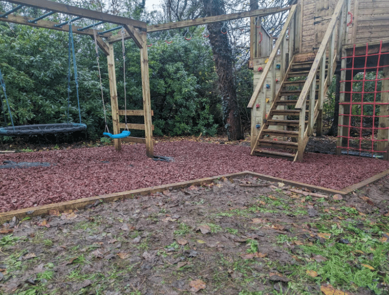 Playground with climbing structure, swings, and wood-chipped ground surrounded by trees.