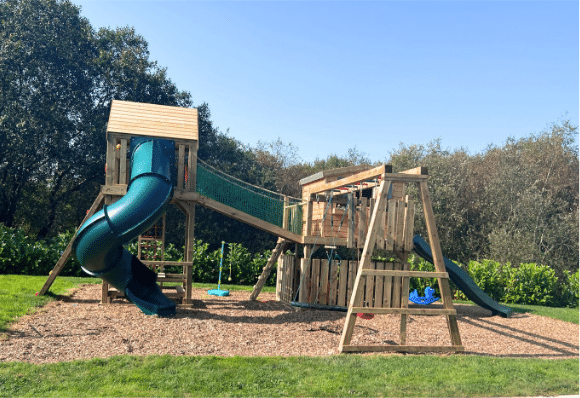 Wooden playground set with slides, swings, and climbing areas on a grassy field under a clear blue sky.