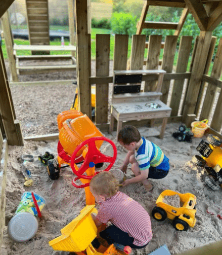 Two children playing in a wooden sandbox with toy trucks and sand equipment.