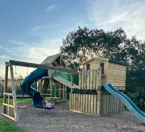 Wooden playground with slides and swings, surrounded by trees under a blue sky.