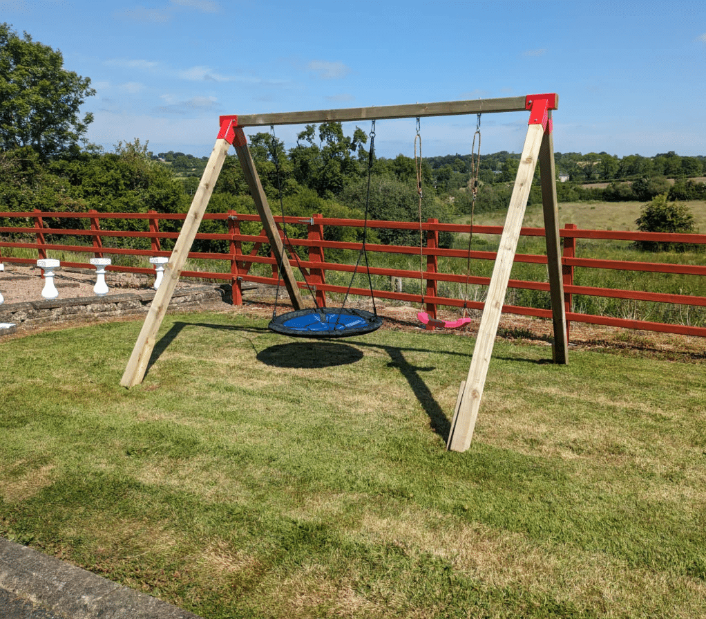 Wooden swing set with two swings on a grassy lawn, surrounded by a wooden fence and trees in the background.