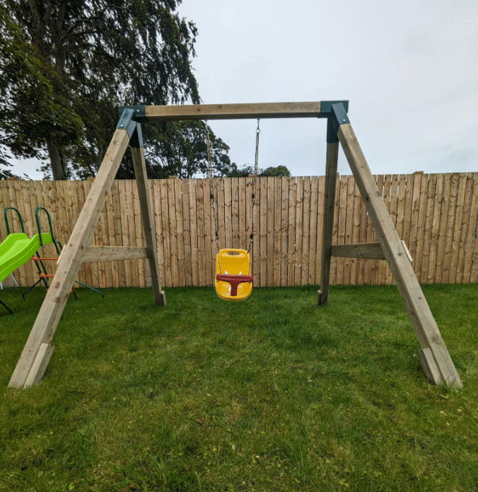 Wooden swing set with a yellow toddler swing seat on a grassy lawn, fenced by wooden panels.