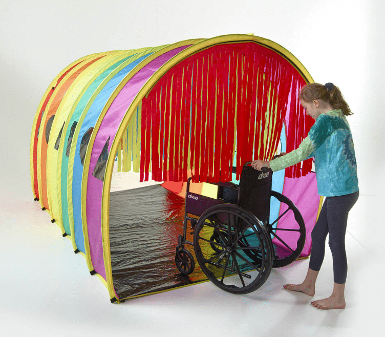 Child pushing a wheelchair through a colorful sensory tunnel with hanging fabric strips.