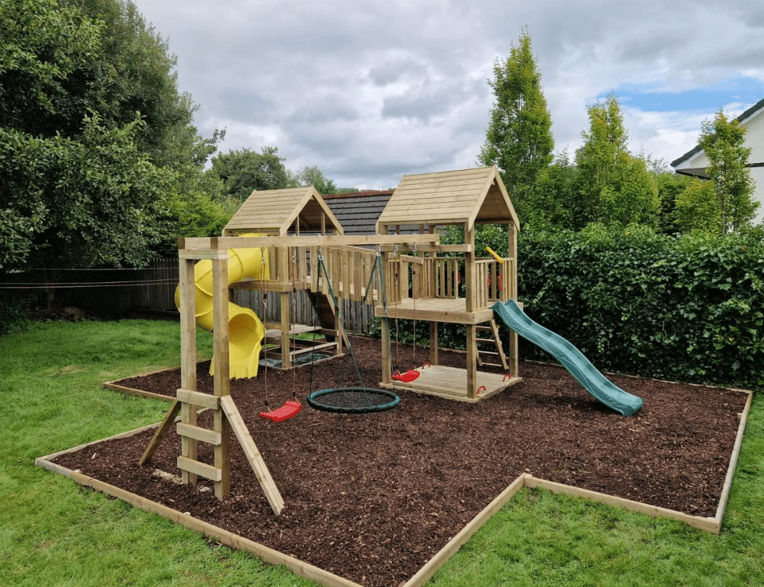 Wooden playground set with slides, swings, and climbing areas in a grassy backyard.
