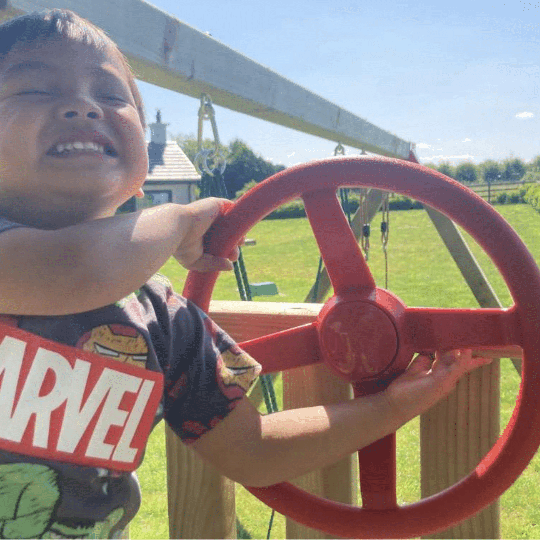 Child smiling while playing with a red toy steering wheel outdoors.