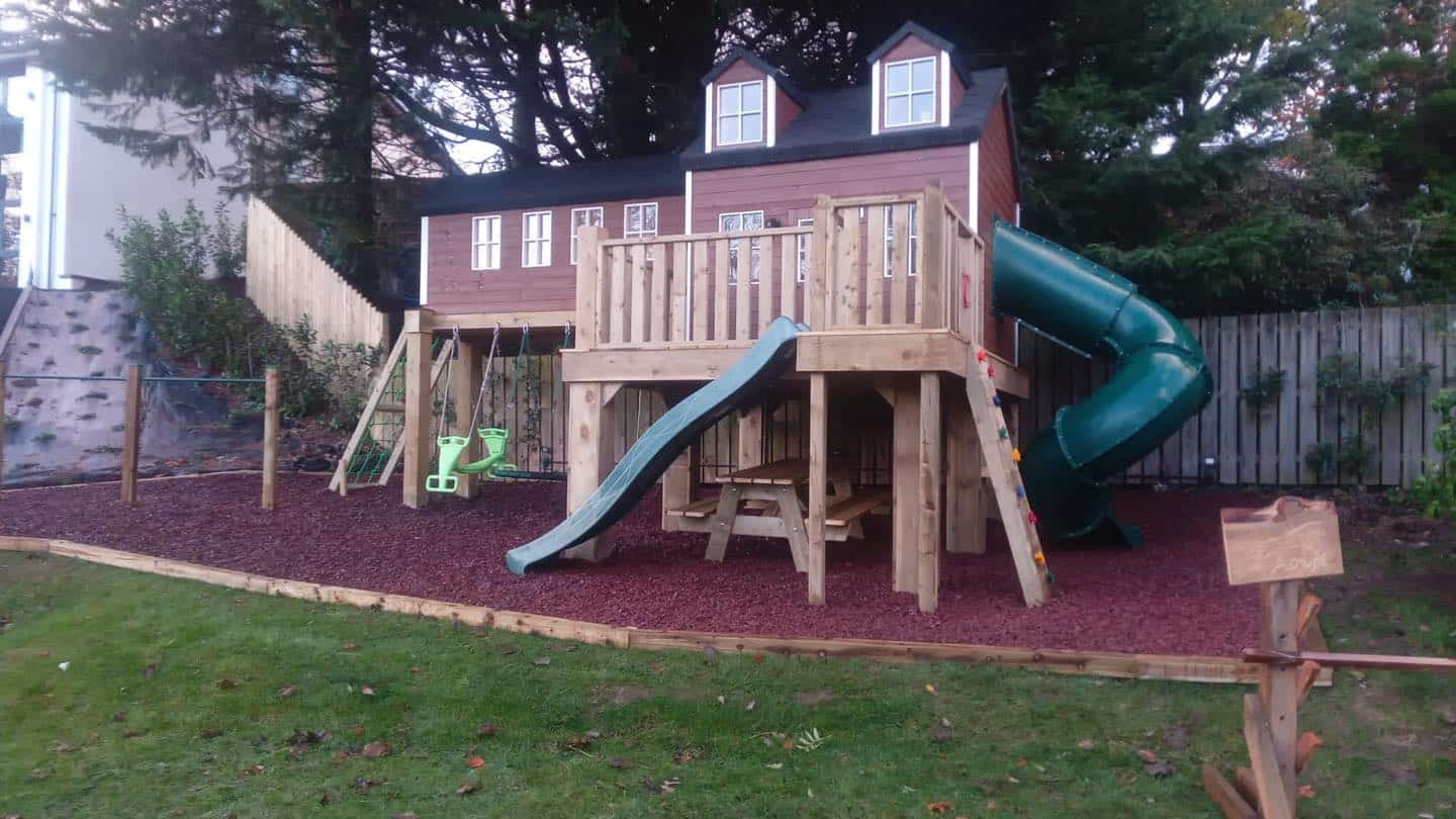 Two-storey playhouse with slides, swings, and picnic table in a backyard setting.