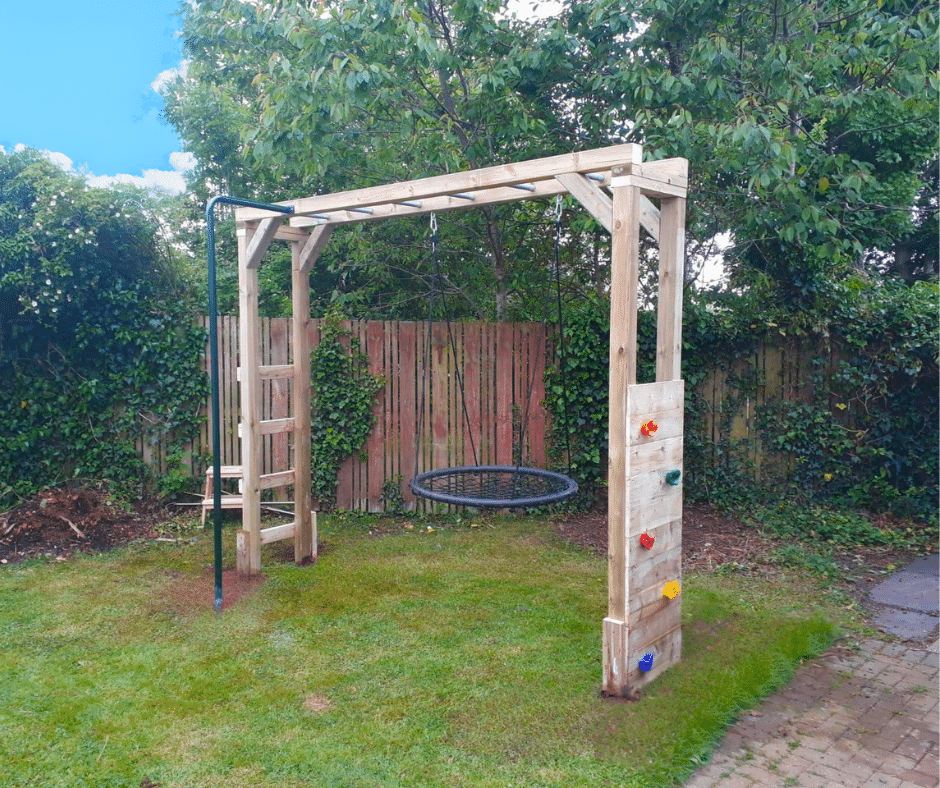 Wooden play structure with rope ladder, climbing holds, and swing in a backyard garden.