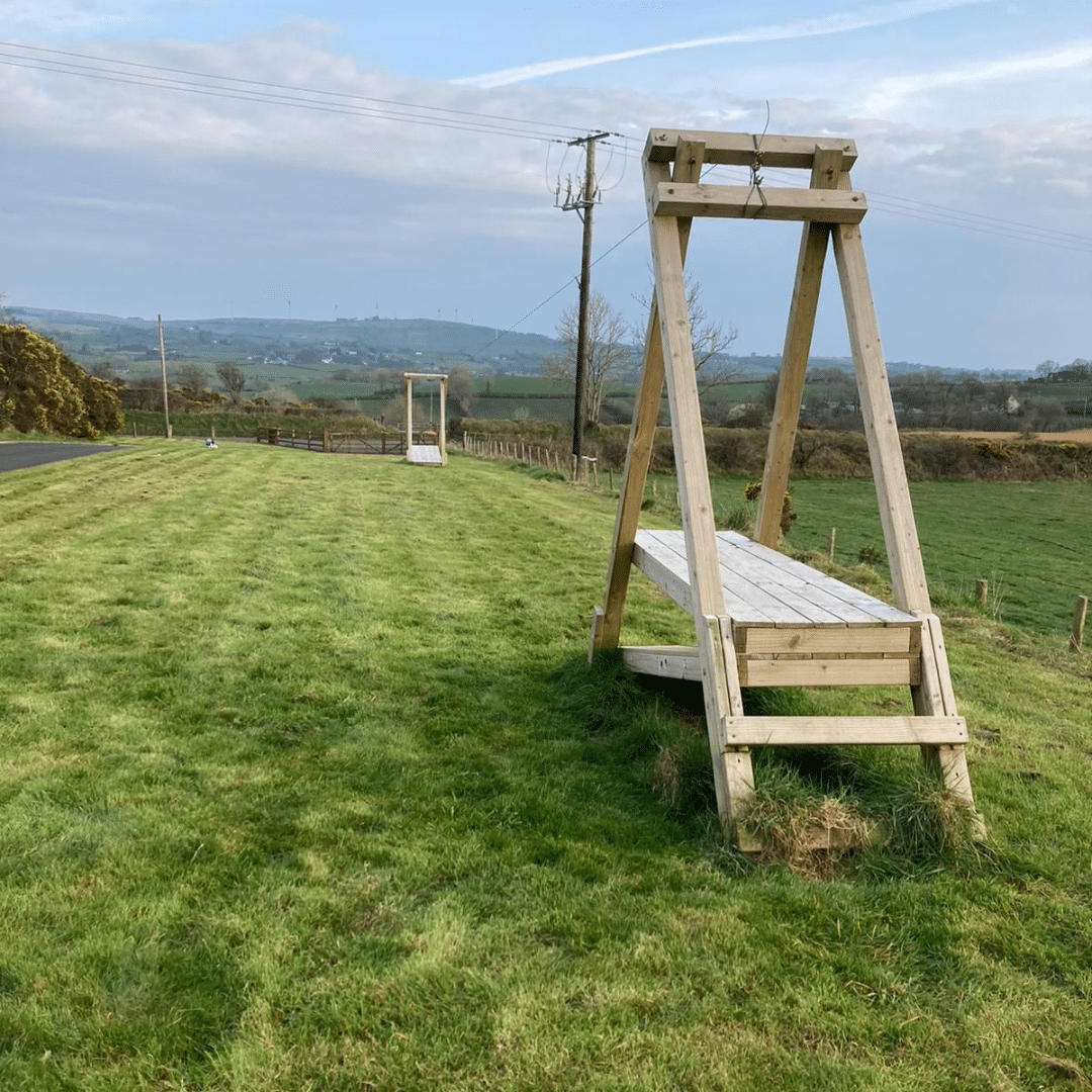Wooden zip line structure in a grassy field with distant hills and a clear sky.
