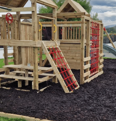 Wooden outdoor playground structure with climbing nets and rings, surrounded by mulch.