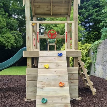 Children's wooden play structure with a climbing wall and green slide in a backyard setting.