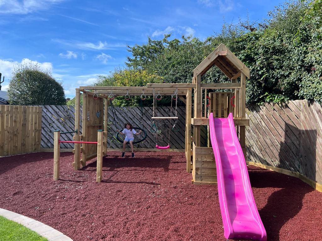 Child playing on wooden playset with pink slide in sunny backyard.