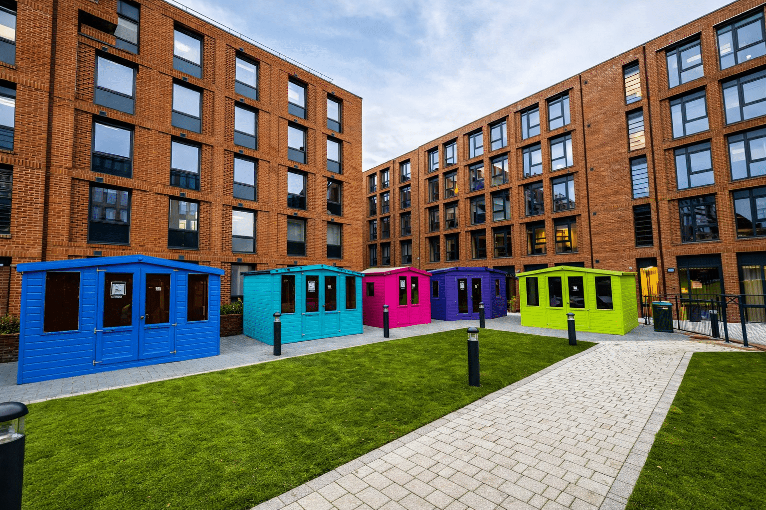 Colorful huts with bright walls outside brick buildings on a grassy pathway.