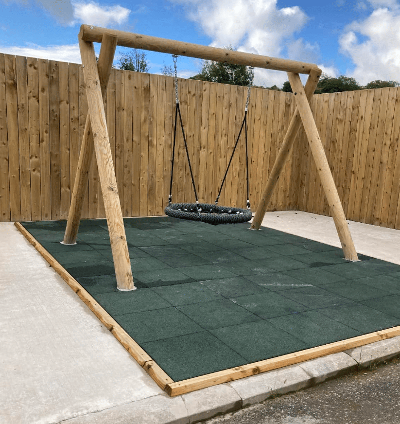 Children's swing set on safety tiles in a fenced outdoor area.
