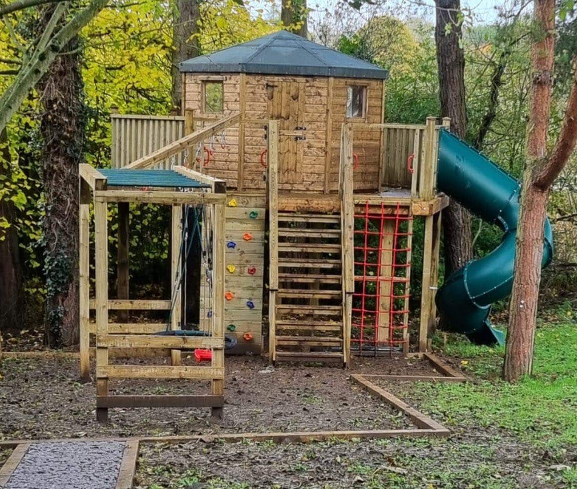 Wooden treehouse with slide, climbing wall, and swing set in a forested area.