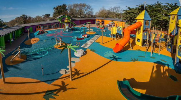 Colorful outdoor playground with slides, climbing structures, and water-themed decorations under a clear sky.